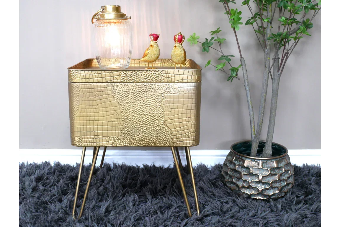 Gold metal side table with textured finish, lamp, and ornaments on top beside potted faux tree