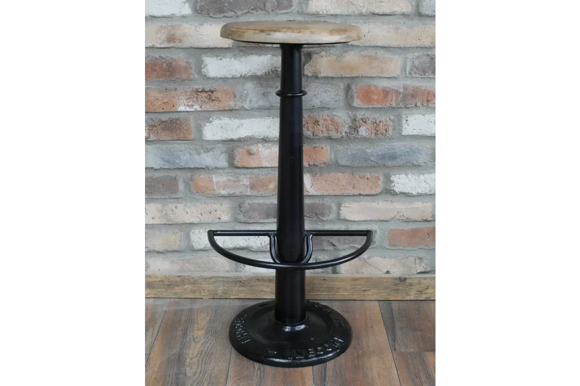 Industrial bar stool with round wood seat, black metal base, and footrest against brick wall