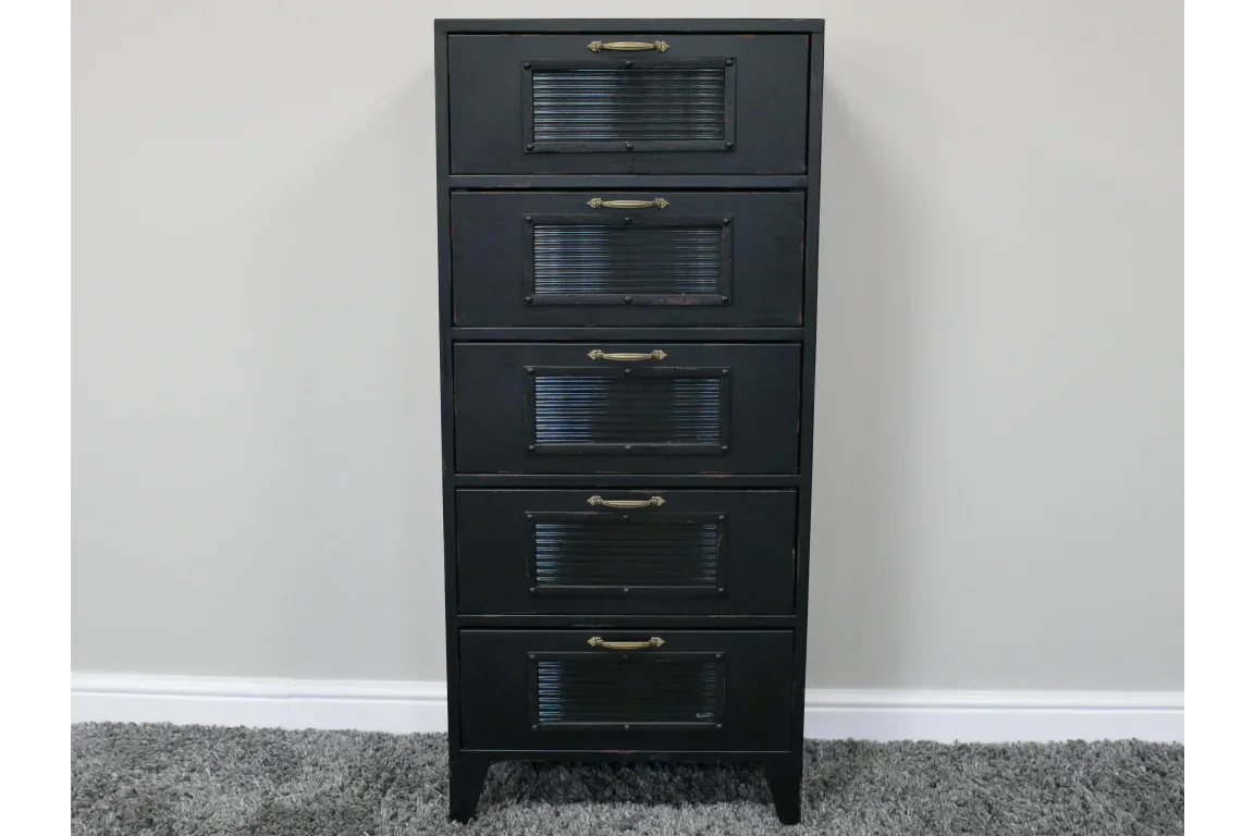 Tall black chest of drawers with metal handles and ribbed front panels on grey carpet