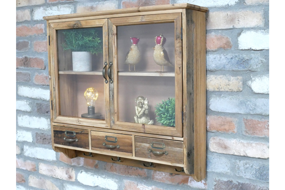 Wooden wall unit with glass doors, three small drawers, and metal handles, shown at slight left angle on brick wall.