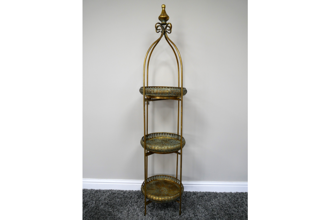 Tall 3 tier metal shelves with decorative round trays in gold and green, standing on a carpeted floor against a plain wall.