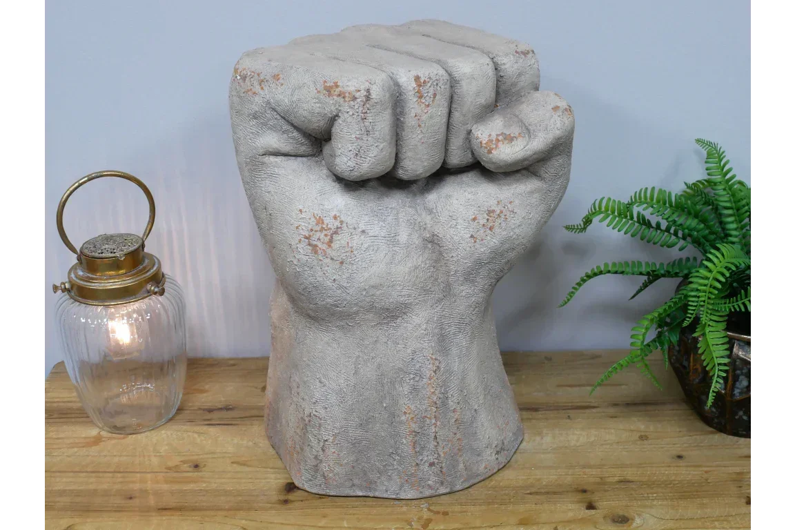 Concrete fist sculpture ornament on wooden table beside glass lantern and potted fern