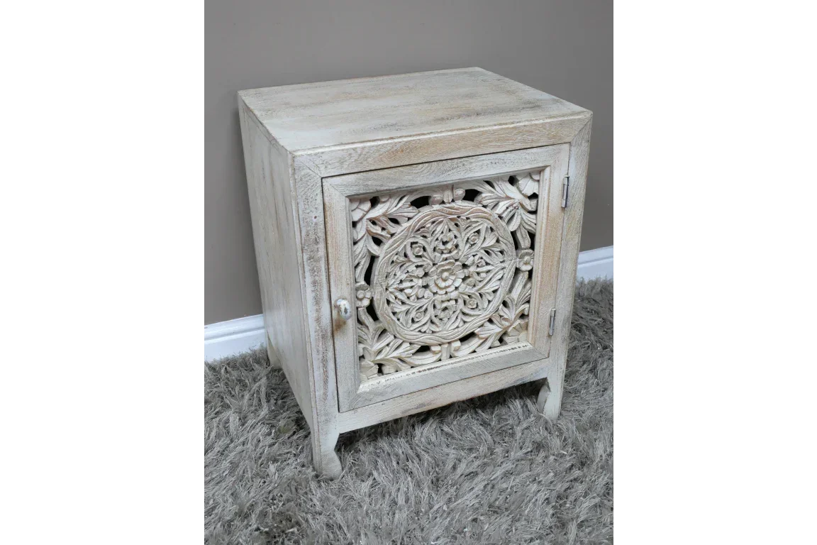 Whitewashed wooden bedside cabinet with carved floral door detail on textured rug