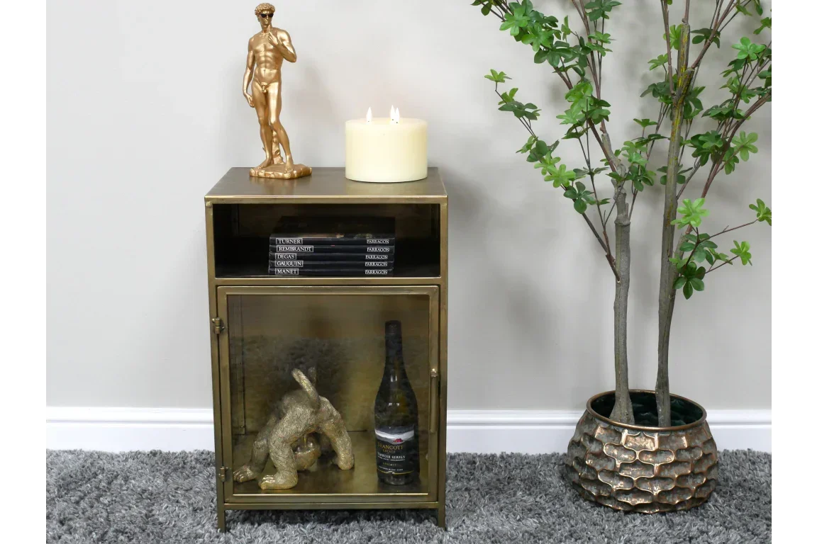 Brass side cabinet with glass door, sculpture, candle, and bookshelf, beside green plant