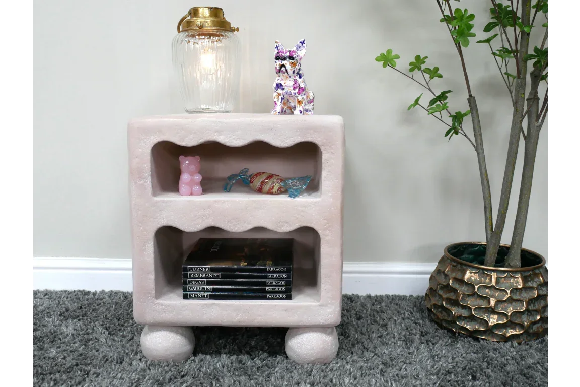 Textured pink bedside table with shelves, lamp, decor objects, and books on grey carpet