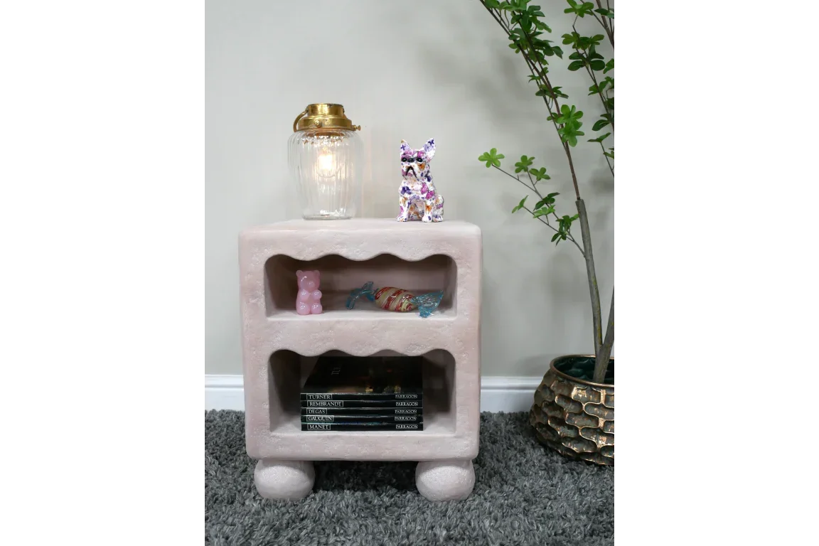 Pink textured bedside table with rounded legs, open shelves, lamp, and decor accents