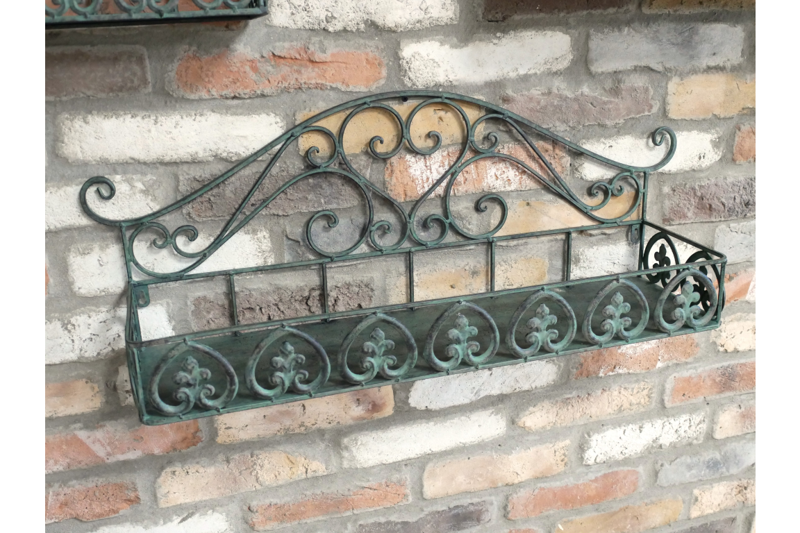 Greenish-blue metal wall shelf with ornate scrollwork, mounted at an angle on a brick wall background.