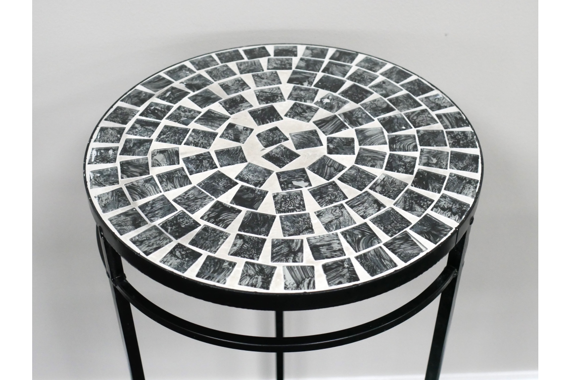 Round side table with black and white mosaic tile top and black metal legs, angled view against white wall.