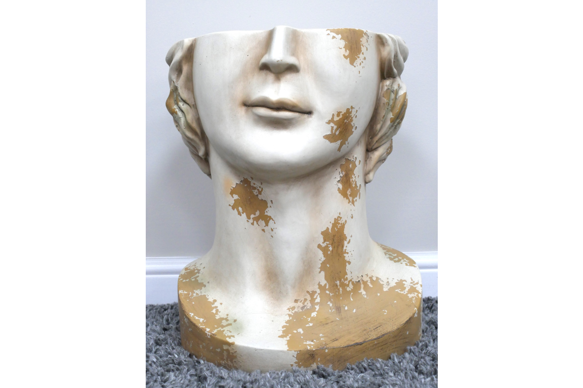 White and gold distressed ceramic head-shaped side table on gray carpet with white baseboard background