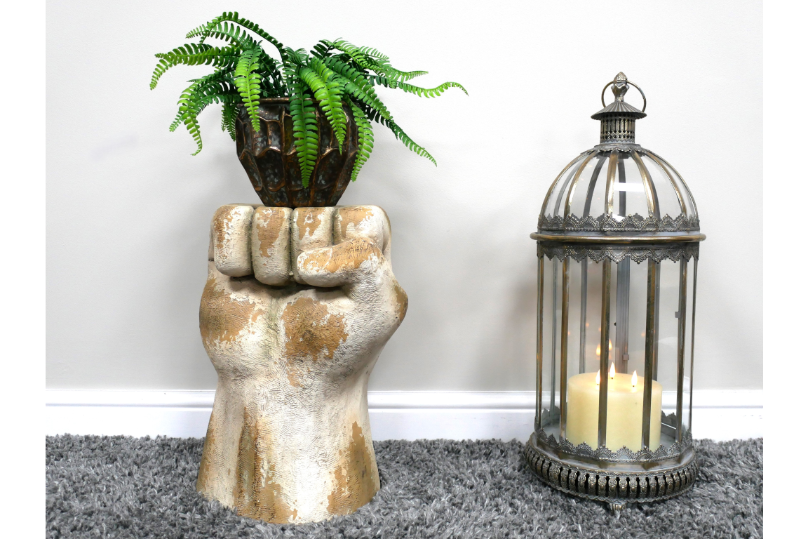 Side table shaped like a raised fist in distressed white and gold finish with a potted fern on top, next to a decorative