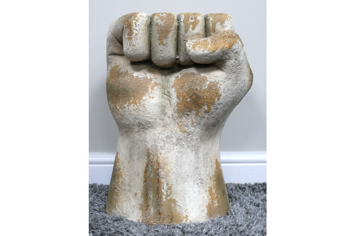 Side table shaped like a clenched fist, distressed white and beige finish, textured surface, standing on gray carpet.