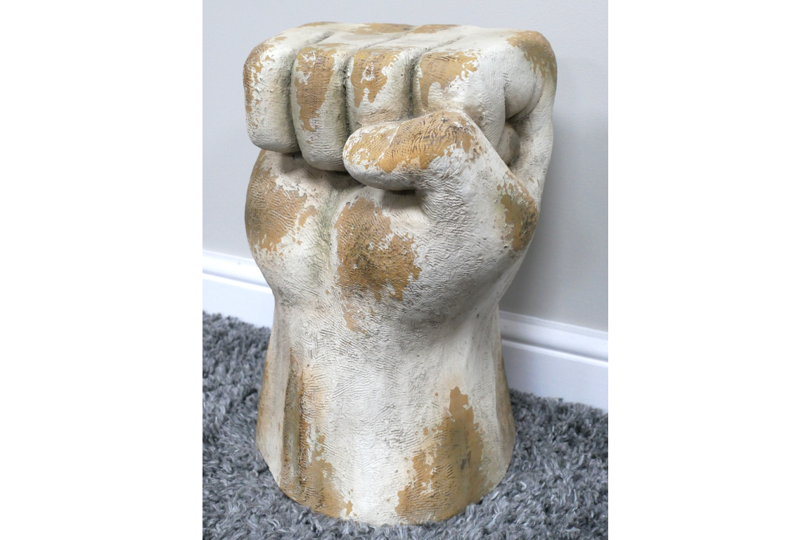 Side table shaped like a clenched fist, weathered white and beige finish, displayed on gray carpet near white wall.