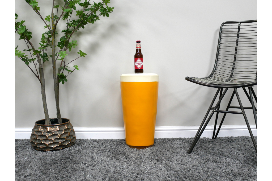 Round yellow side table with white top, ceramic material, shown with bottle on top, next to black wire chair and potted