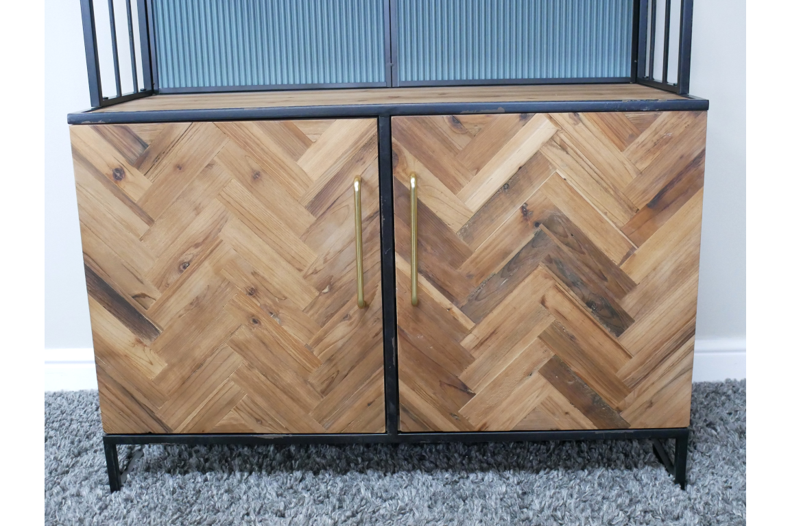 Bar cabinet with herringbone patterned wood doors, black metal frame, gold vertical handles, shown front view on gray carpet.