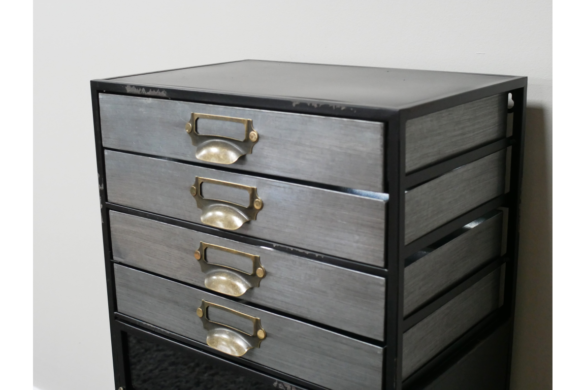 Industrial drawer cabinet with four metal drawers and brass handles in black metal frame, angled view against white wall.