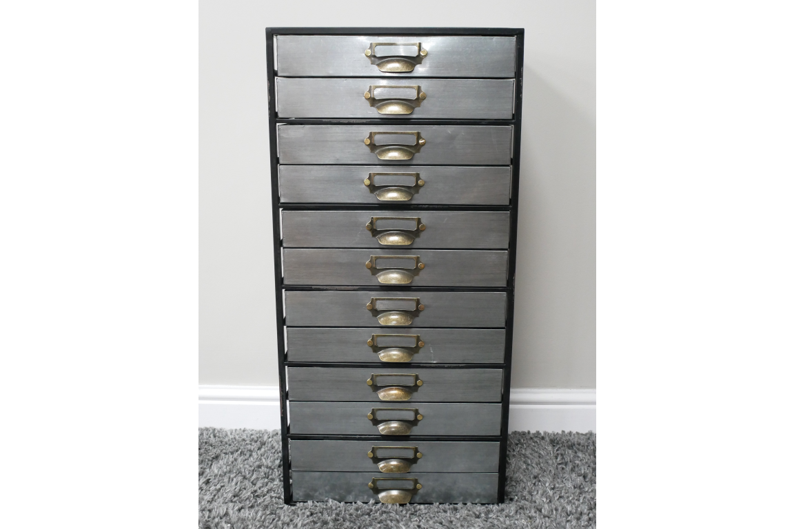 Tall industrial drawer cabinet with ten grey metal drawers and brass handles, standing on grey carpet against white wall.