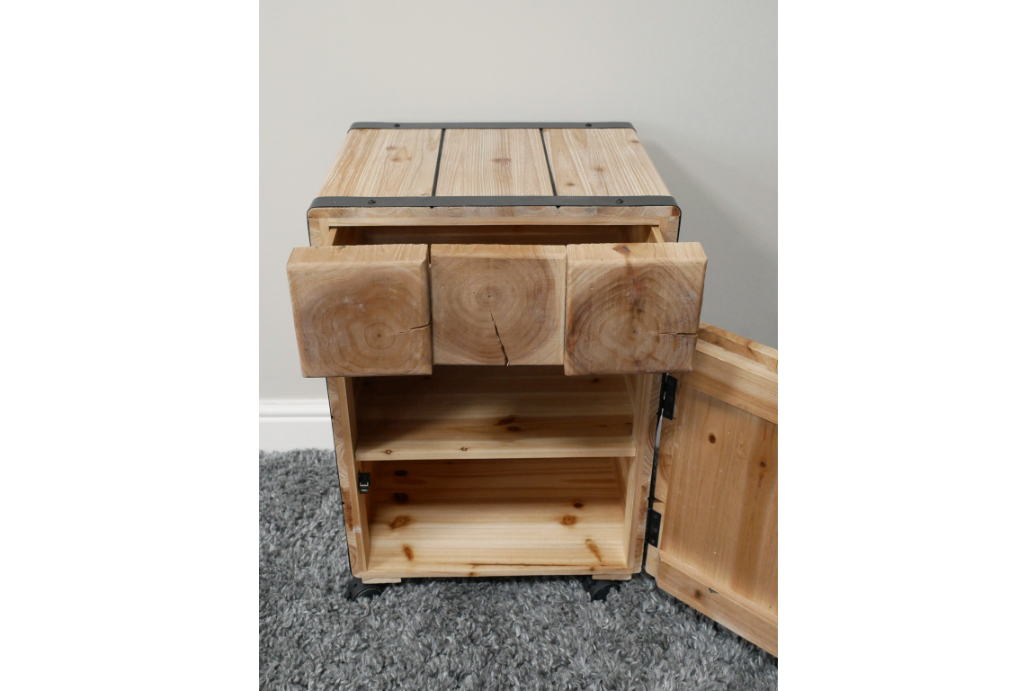 Light wood chunky bedside cabinet with open drawer and door showing interior shelving, placed on gray carpet.