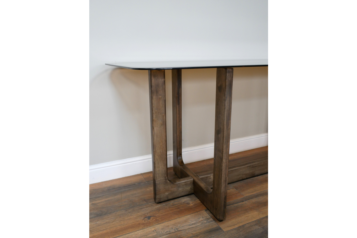 Dining table with clear glass top and wooden legs, side angle view on wooden floor against white wall.