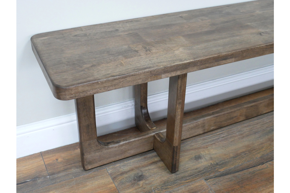 Long wooden bench with sturdy legs, medium brown finish, angled side view on wooden floor against white wall.