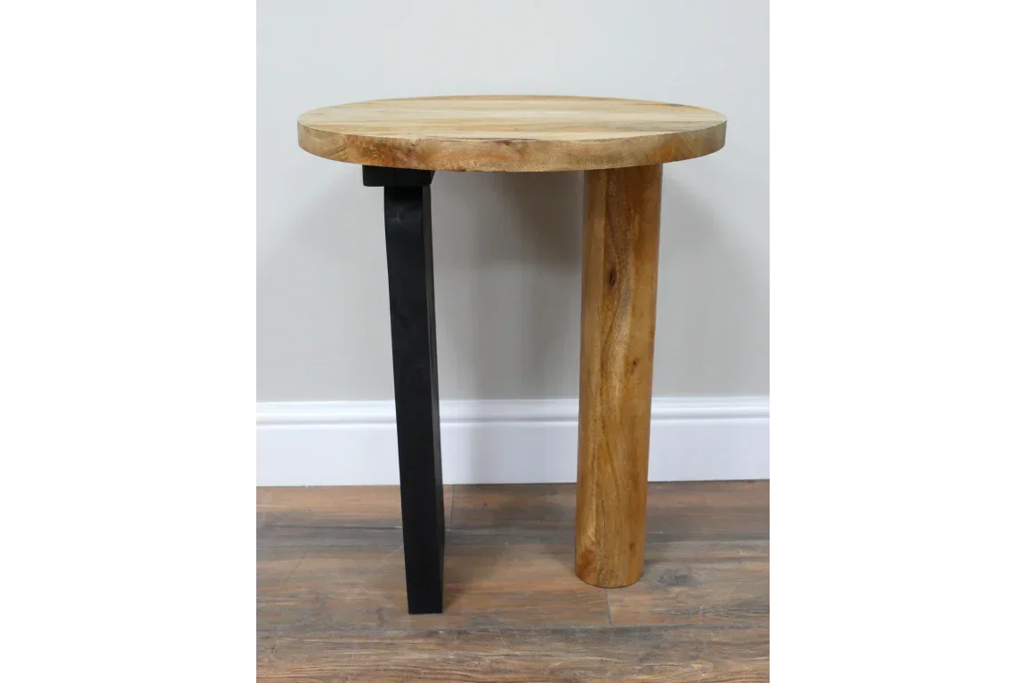 Round wood side table with one black metal leg and one wooden leg on laminate floor