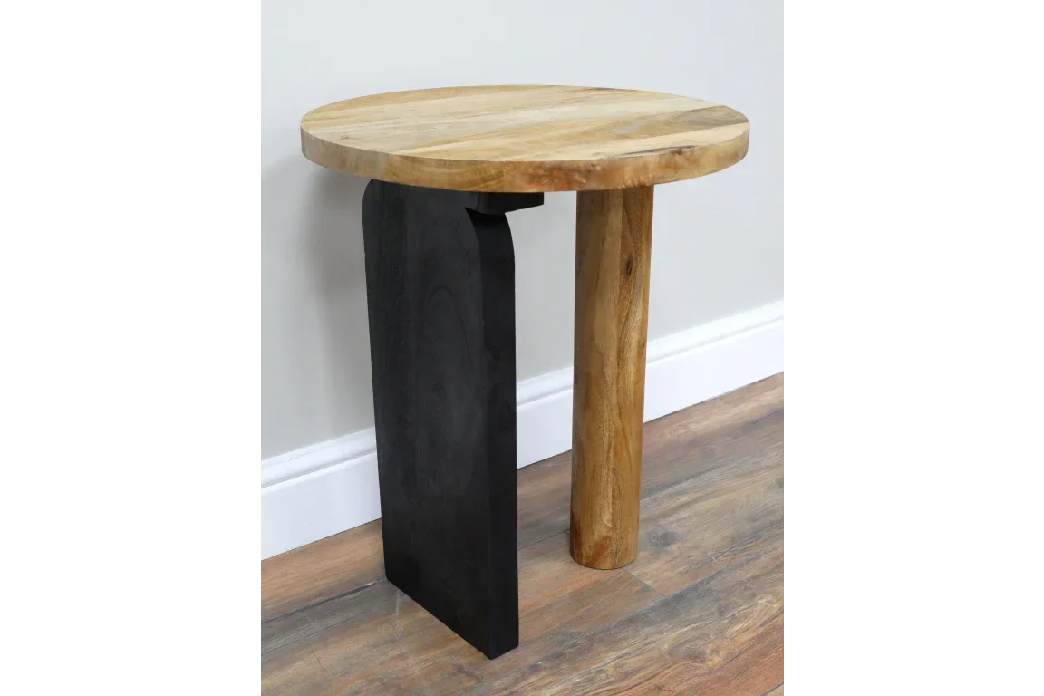 Round wood side table with one black leg and one natural wood leg on laminate floor