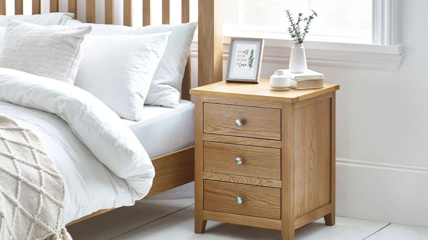 Oak bedside table with three drawers beside bed, styled with books, vase, and framed print