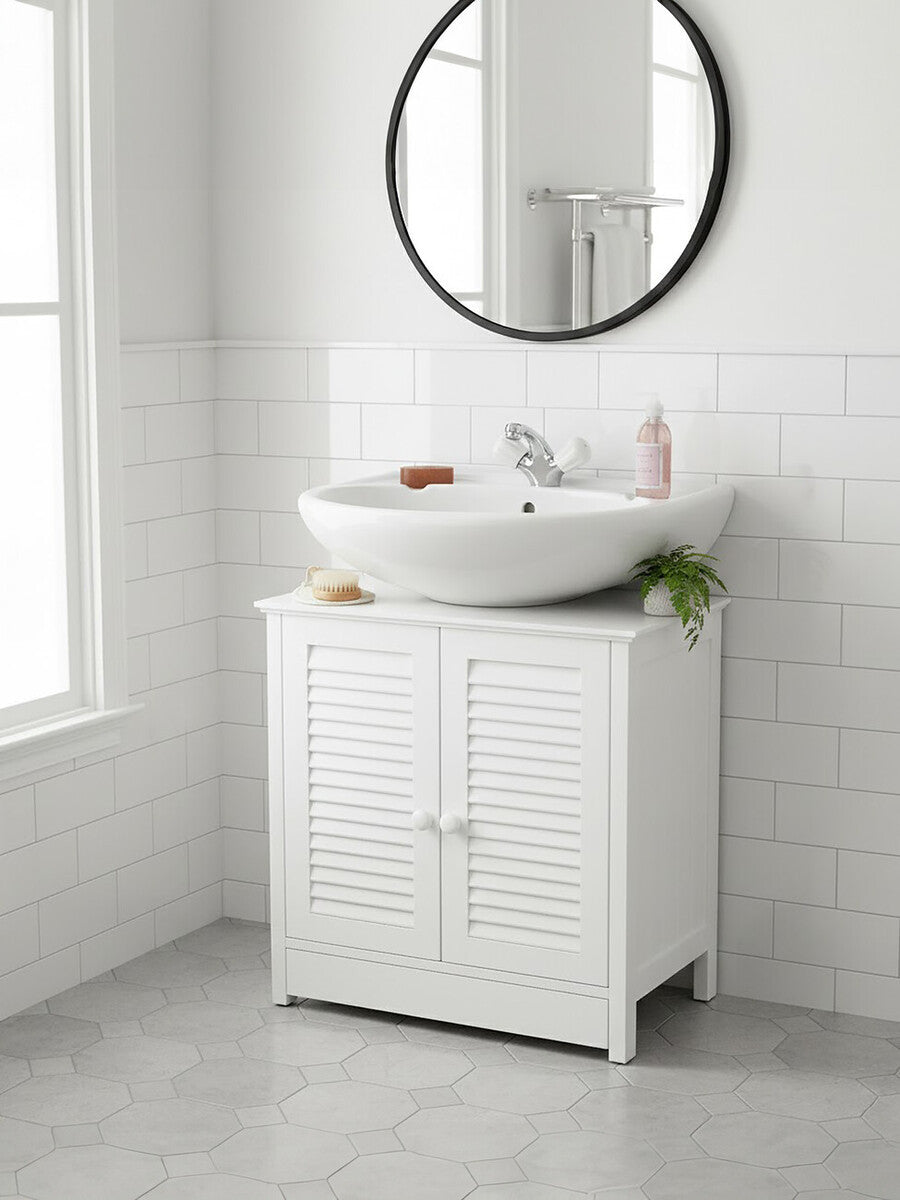 White wood under sink bathroom cabinet with shutter-style doors, a round mirror above, and a modern sink on top. The cabinet is positioned against a light-colored wall with a window, and a small plant and toiletries are placed on the sink. The floor is tiled in a light gray color