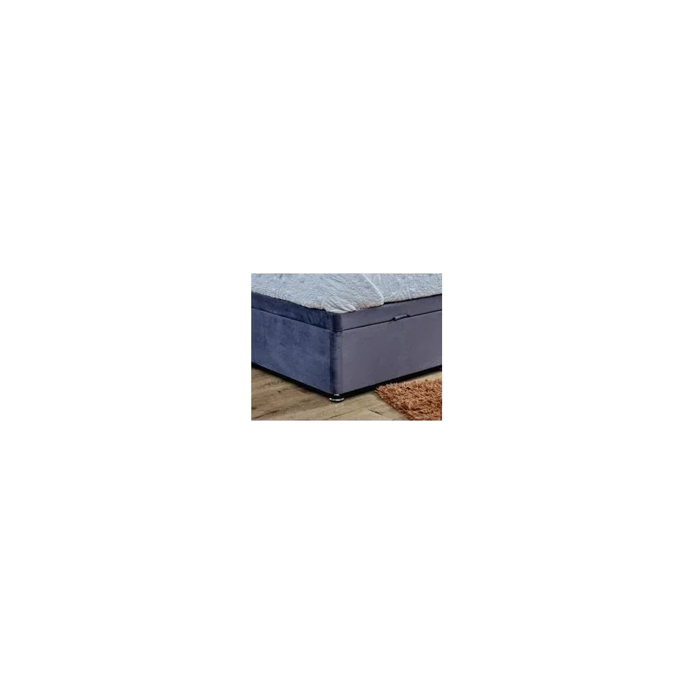 Blue velvet ottoman bed base with storage, shown on wooden floor beside a rug