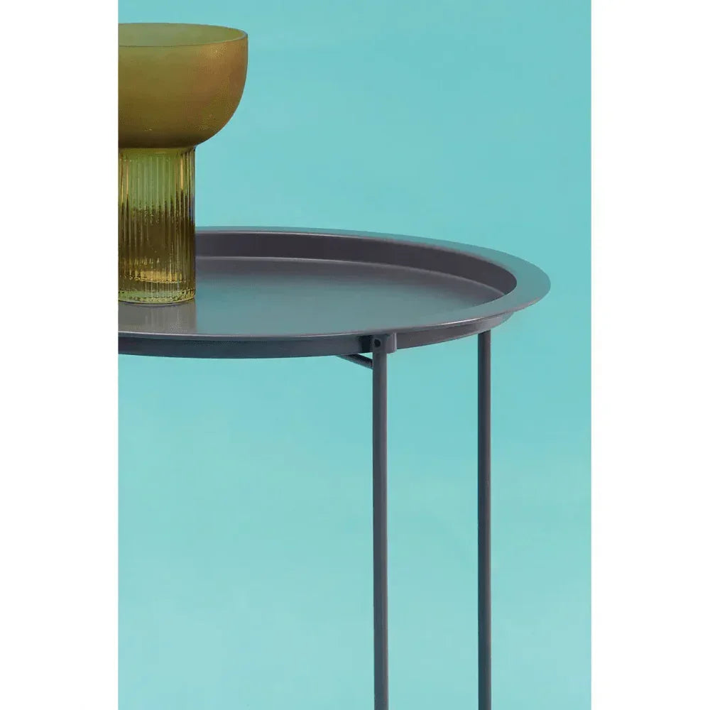 Round metal side table with slim legs and amber glass vase on blue background