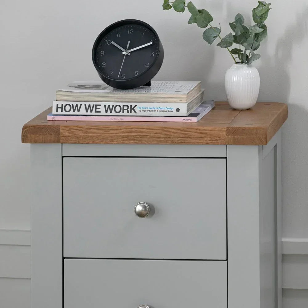 Grey bedside table with oak top, stack of books, black clock, and white vase with greenery