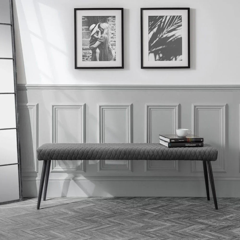 Grey upholstered bench with diamond quilting, angled black legs, books and cup on top