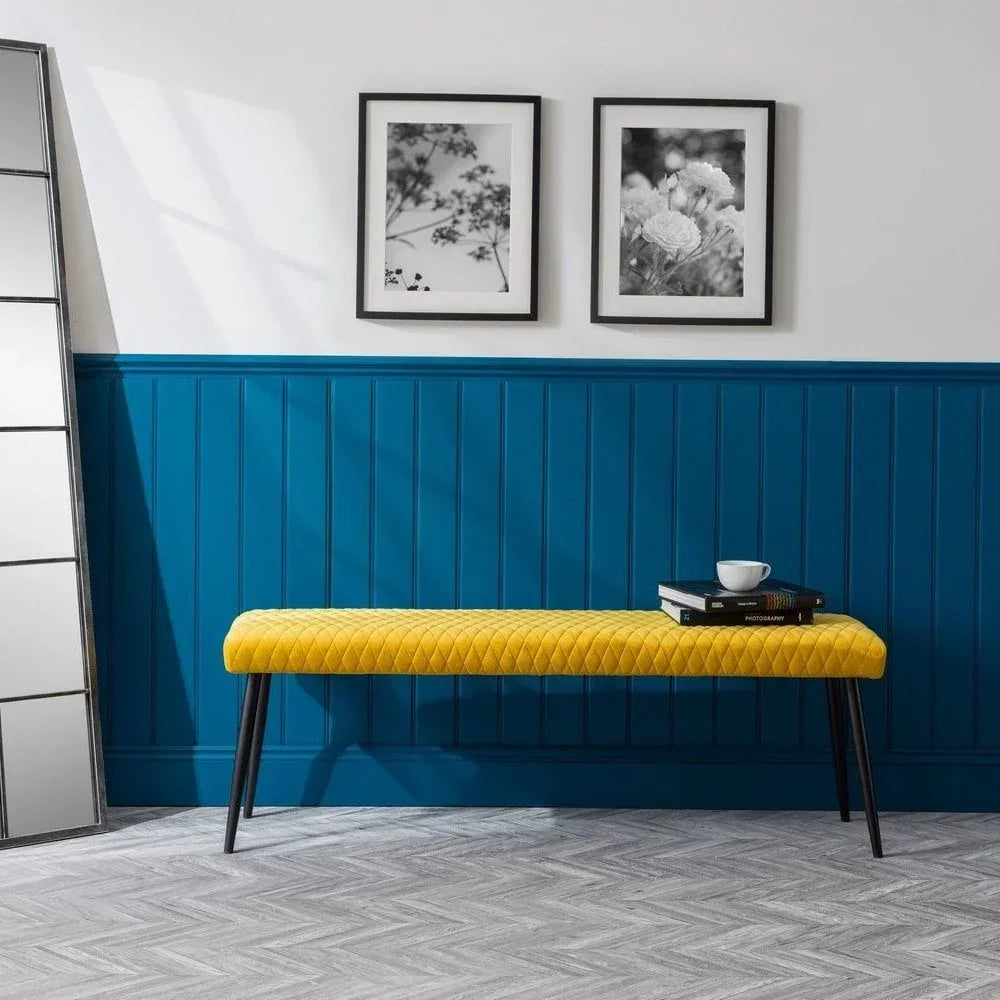 Yellow upholstered bench with black legs beside books and cup against blue wall paneling