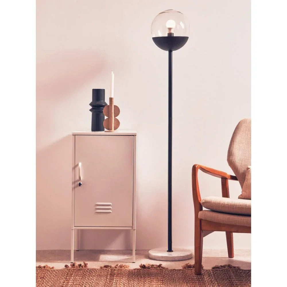 Modern floor lamp with glass globe shade beside beige metal cabinet and wooden armchair