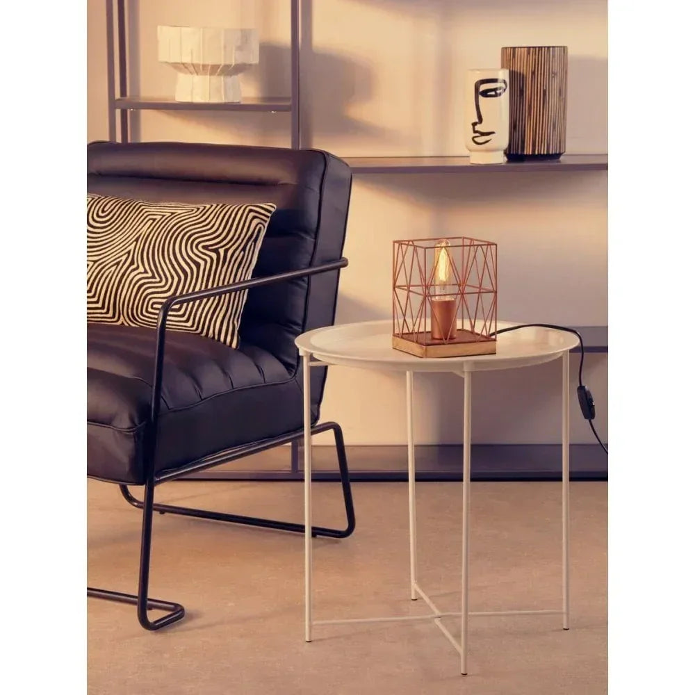 Round white metal side table with modern cage lamp beside black armchair and patterned cushion