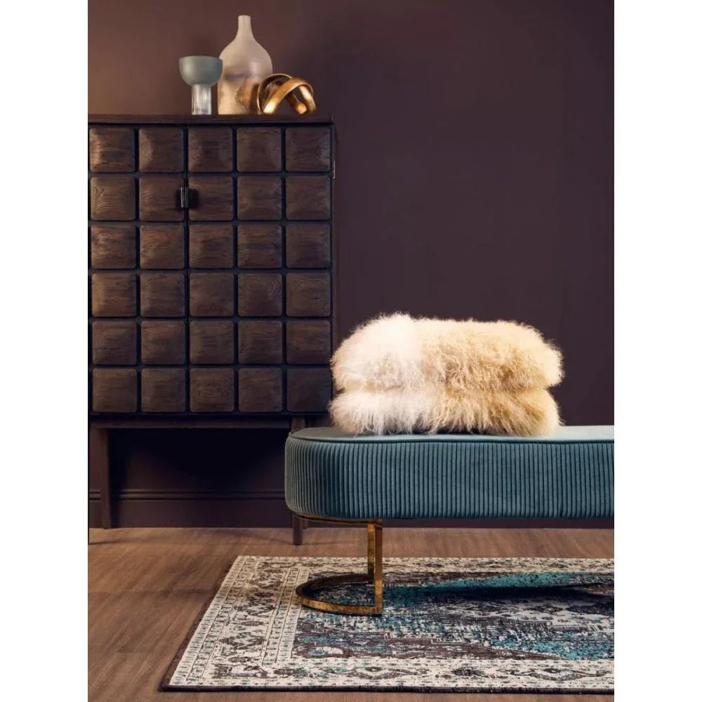 Velvet ottoman bench with gold legs and fluffy throw, beside dark wood cabinet on rug