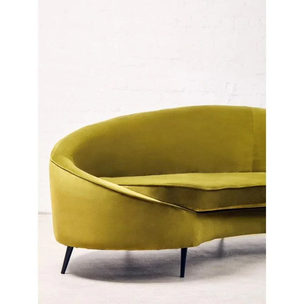 Curved green velvet sofa with slim black legs against a white brick wall background