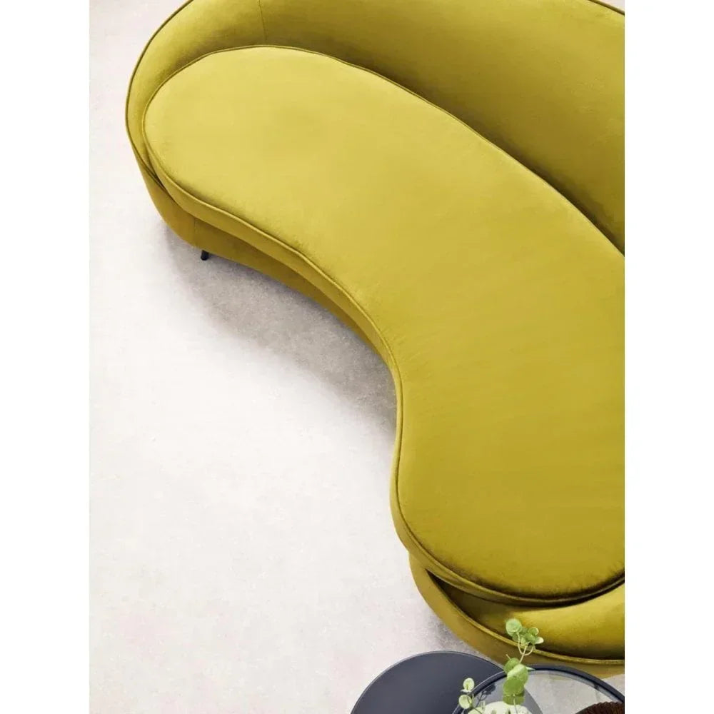 Curved yellow velvet sofa on light carpet with small round black table and plant