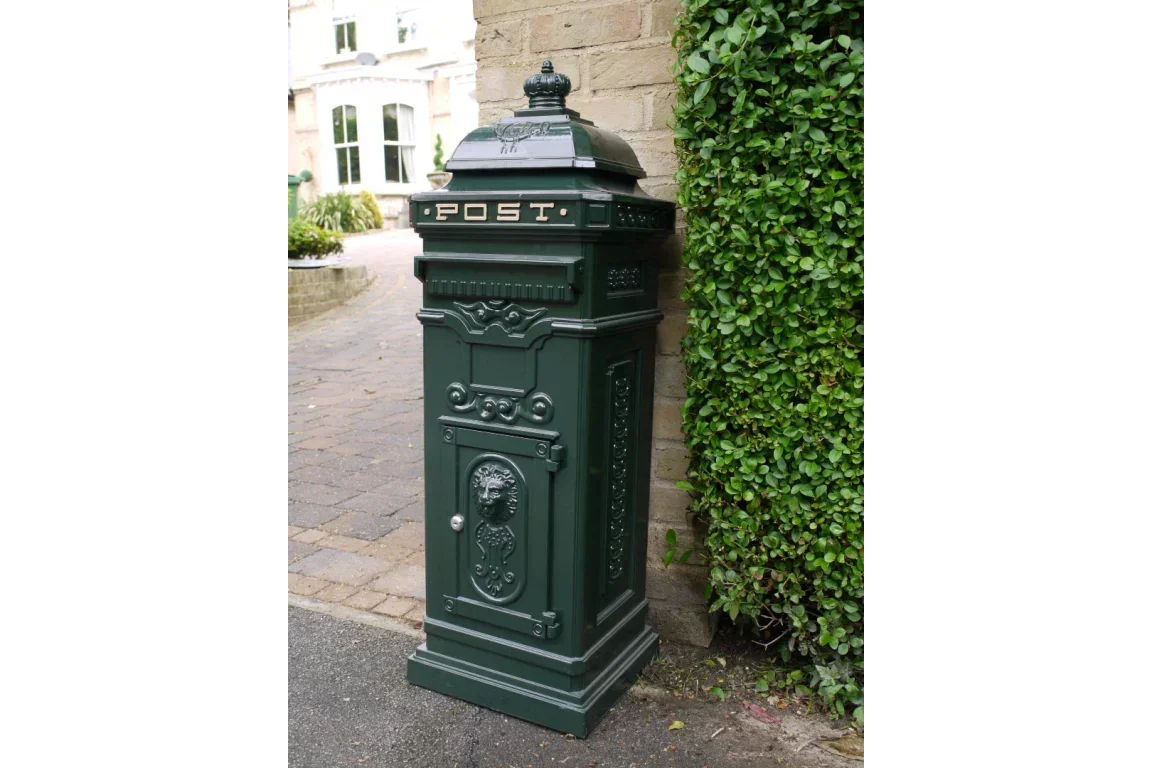 Green cast iron post box with ornate detailing beside brick wall and garden hedge