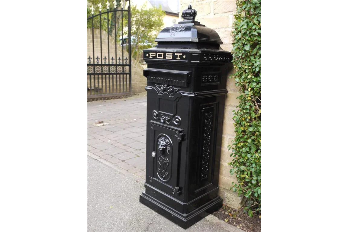 Black cast iron post box with ornate detailing beside stone wall and garden gate