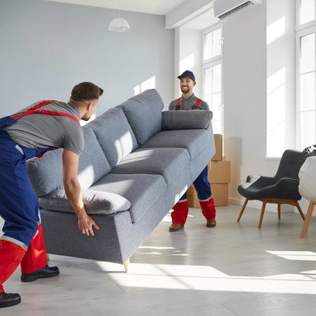 Two movers carry grey fabric sofa in modern living room with armchair and boxes