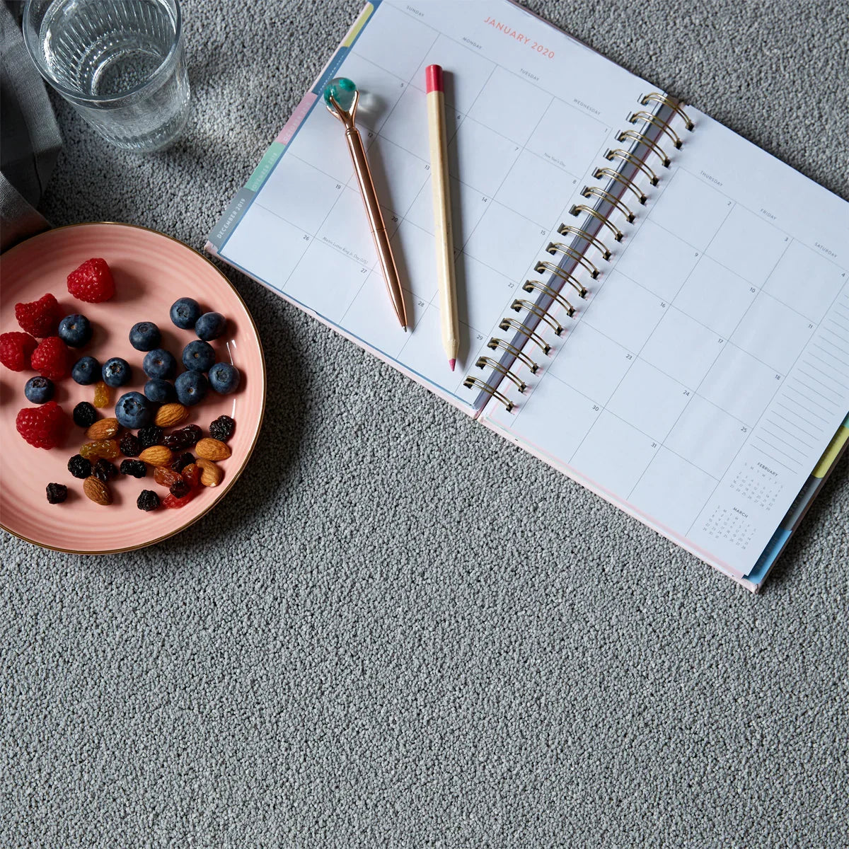 A cozy setup featuring a January 2020 planner with a pen and pencil, accompanied by a plate of fresh berries, nuts, and a glass of water on a soft carpet.