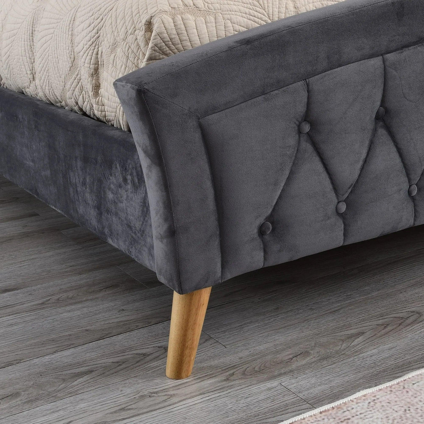 Charcoal upholstered bed frame corner with button tufted detail and wooden leg on grey floor