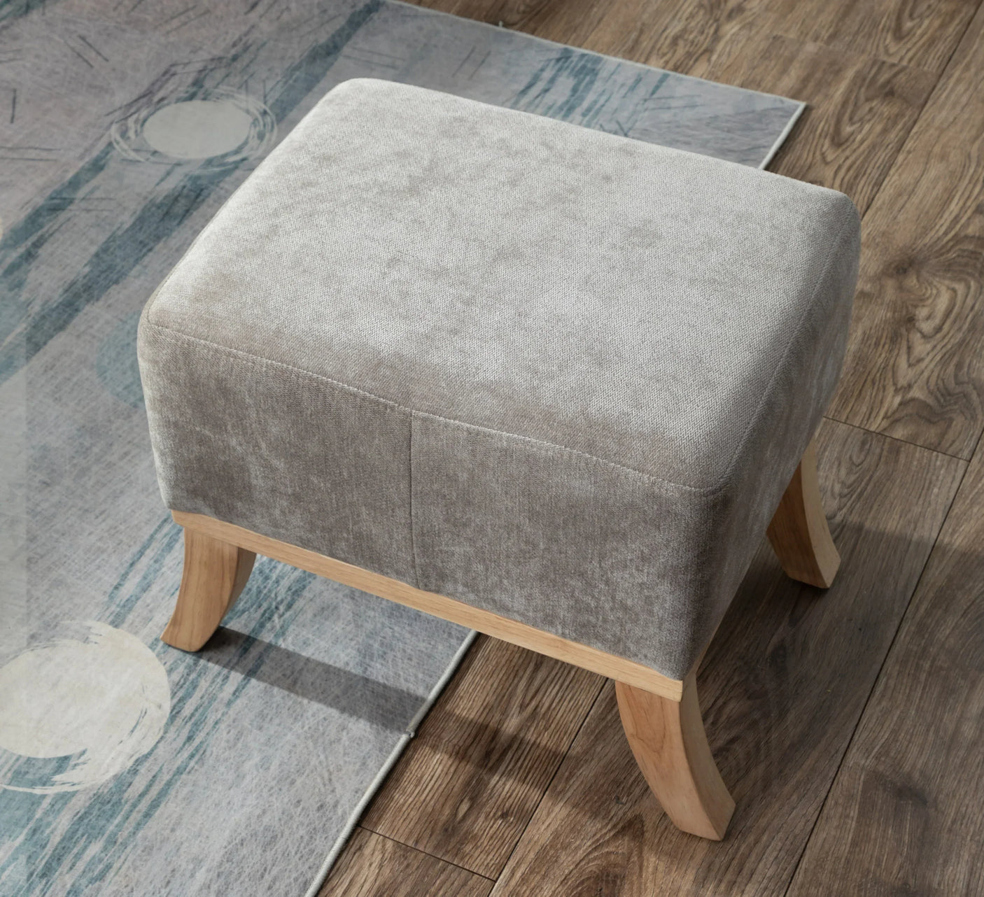Grey fabric footstool with light wooden legs on hardwood floor and patterned rug