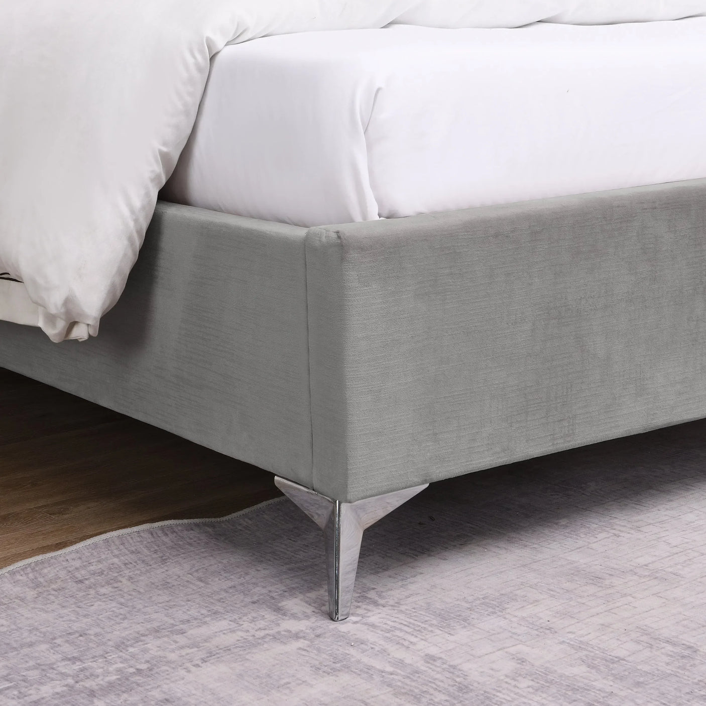 Grey upholstered bed frame corner with chrome legs on light rug and wood floor