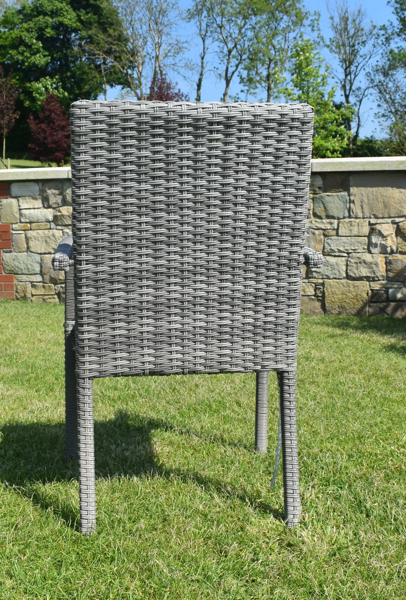 Grey rattan garden dining chair with armrests on lawn, stone wall and trees in background