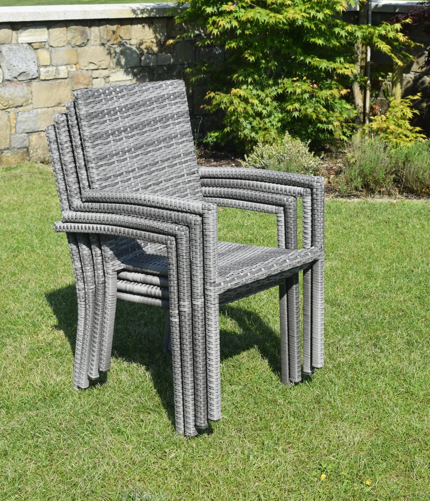 Stacked grey rattan garden chairs on lawn with stone wall background