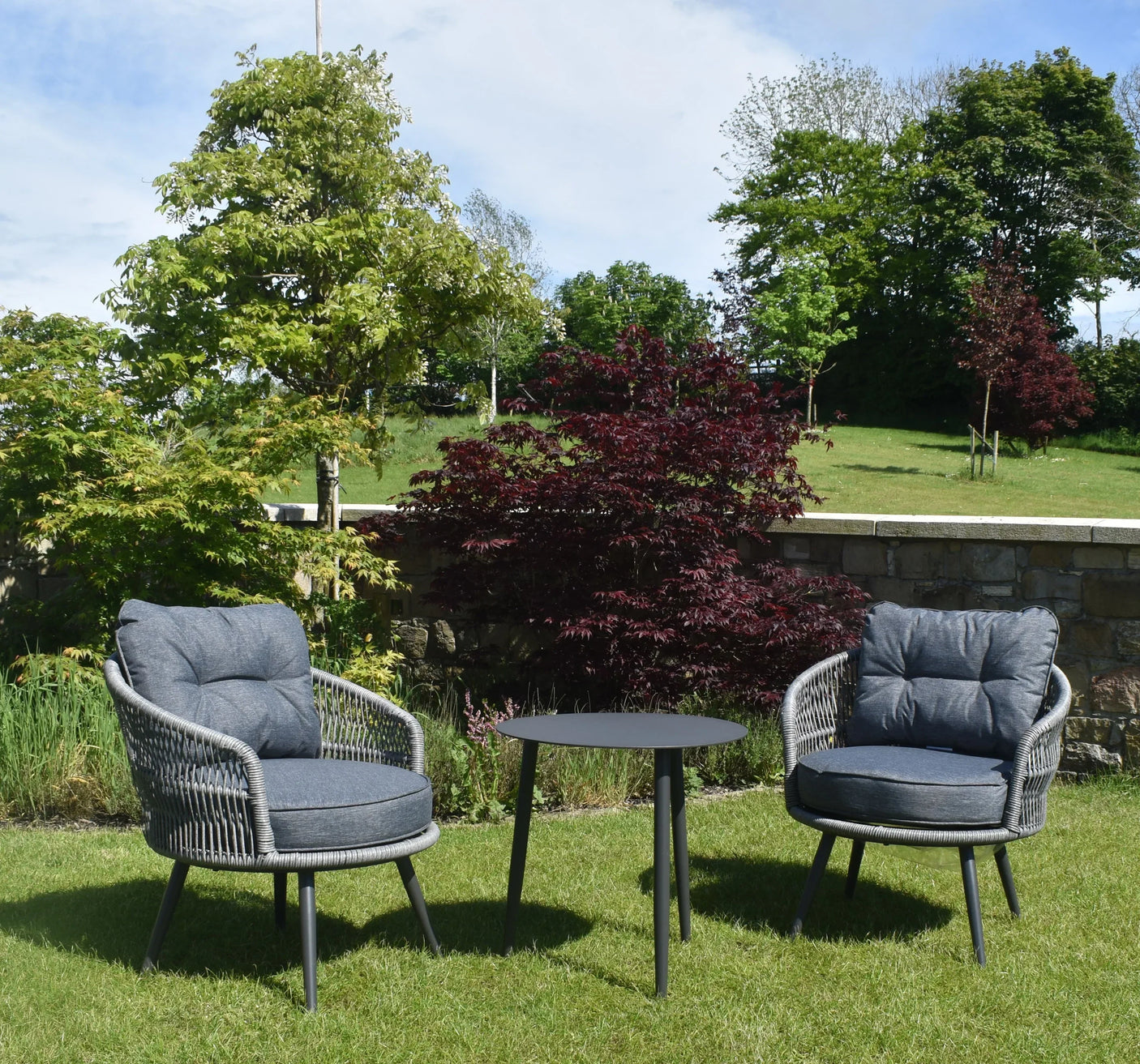 Grey rattan garden chair set with round side table on lawn with trees in background