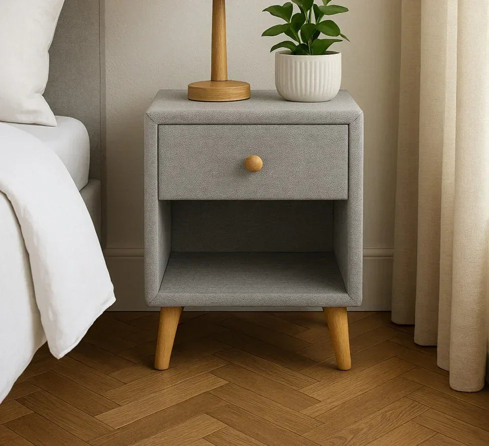 Grey fabric bedside table with wooden legs and drawer beside bed, styled with plant and lamp