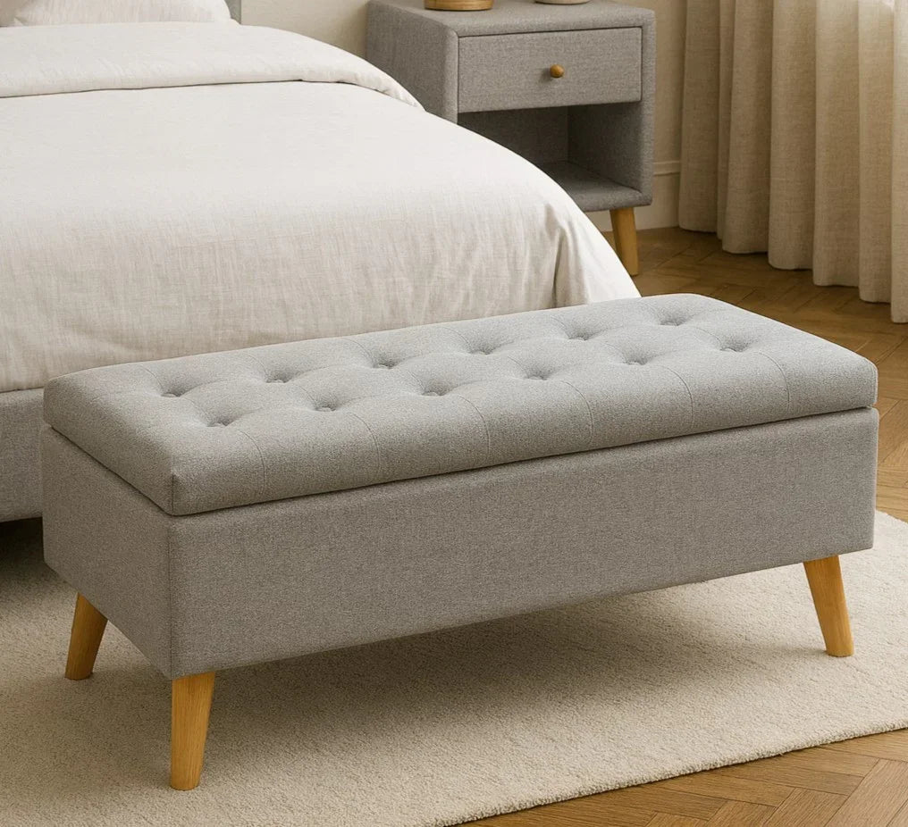 Grey upholstered storage bench with wooden legs at the foot of a bed in a bedroom