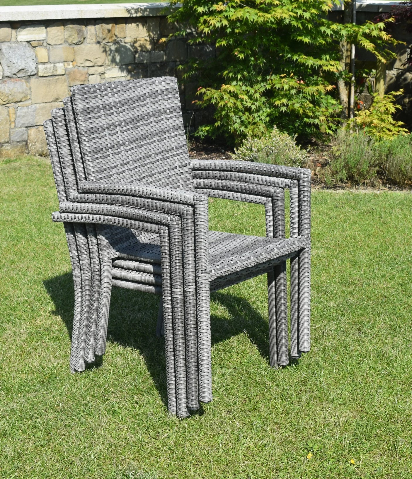 Stacked grey rattan garden chairs on lawn with stone wall and shrubs in background